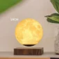 A moon-shaped lamp measuring 14 centimeters in diameter, floating above a wooden base, next to a green plant in a white pot.