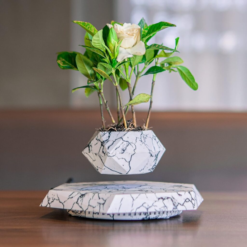 The Levitating Hexagonal Plant Pot features a small green plant with white flowers floating in a white, marbled geometric pot above a matching base, suspended in mid-air through magnetic levitation.