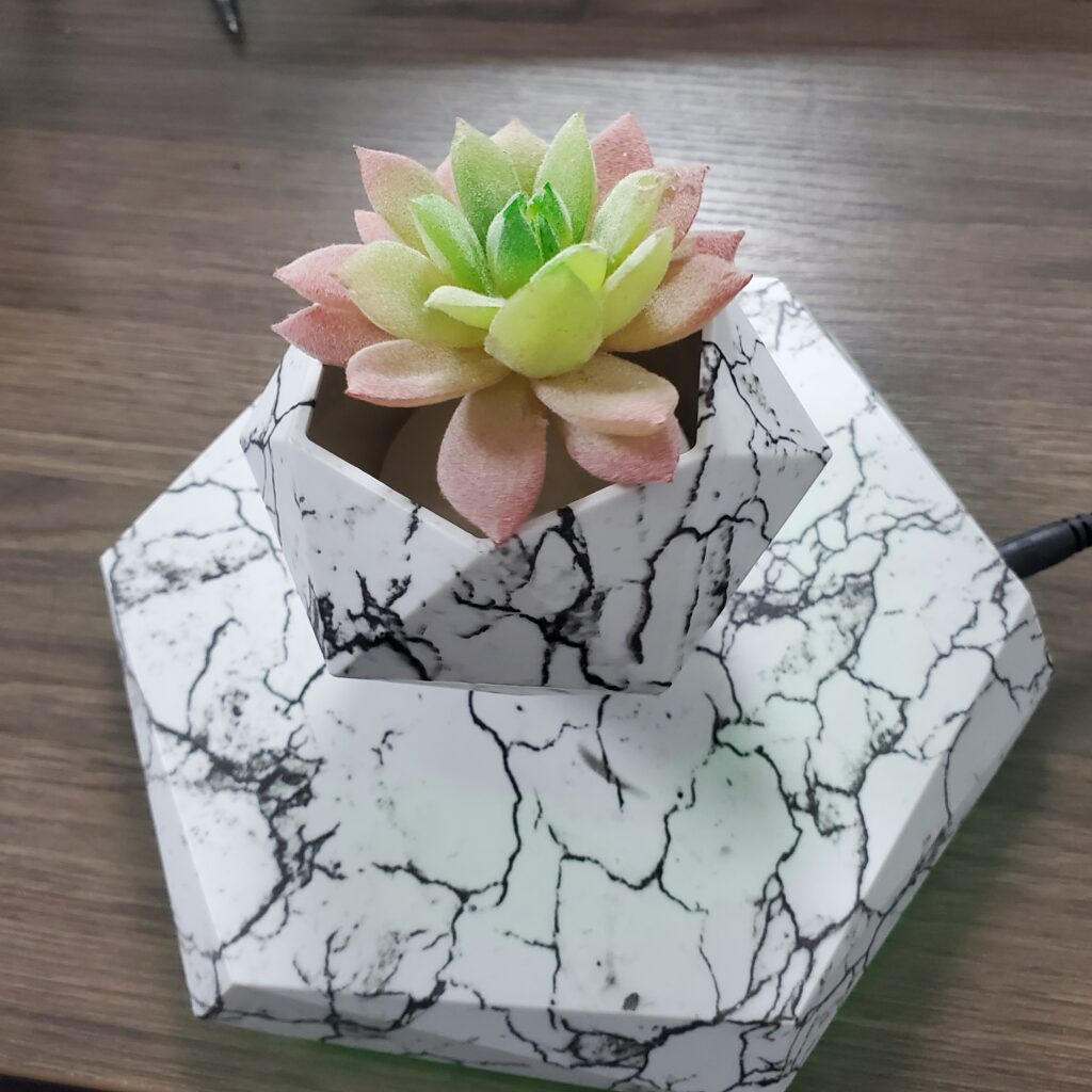 A Levitating Hexagonal Plant Pot featuring a pink and green succulent plant, housed in a geometric white faux-marble planter on a matching base, is placed on a wooden surface.