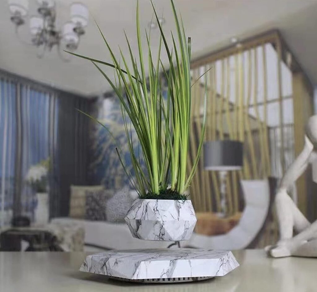 A Levitating Hexagonal Plant Pot with a white, marble-patterned finish and long green leaves floats above its matching base. In the background of the modern living room, you can see a chandelier, blue curtains, and beige furniture.