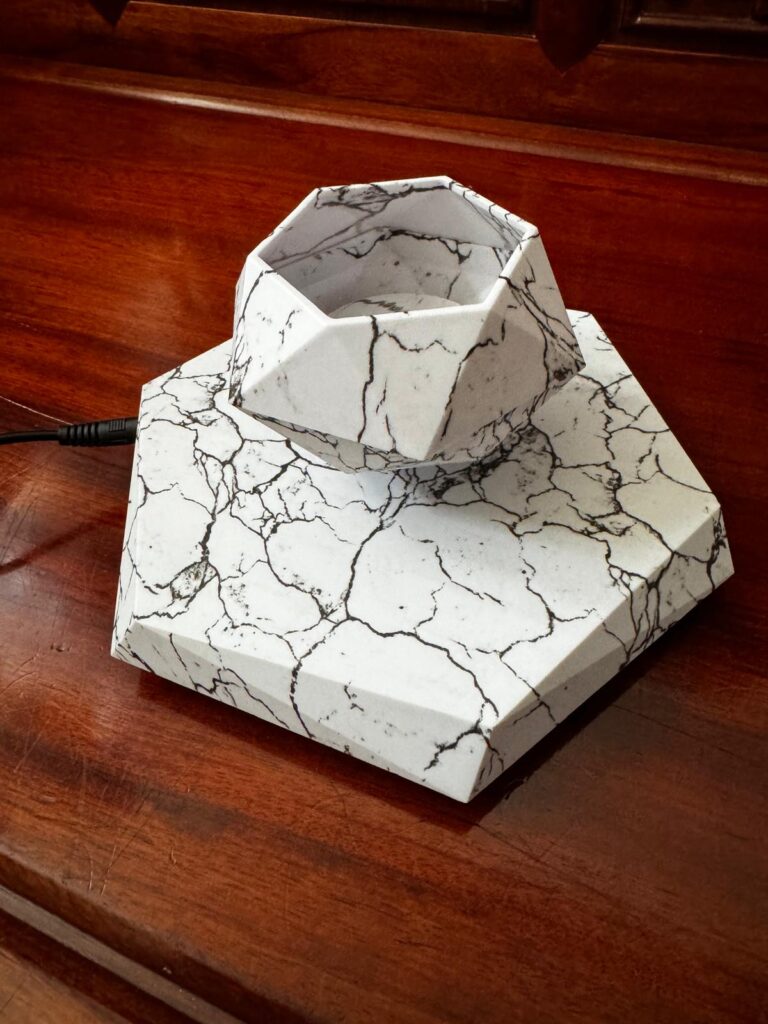 The Levitating Hexagonal Plant Pot, featuring a marble pattern on a wooden base, showcases its multifaceted geometric design with a power cord attached to one side.
