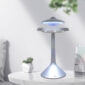 A modern, UFO-shaped Floating UFO Speaker glows with blue light on a white table. A small potted plant and calendar are also on the table, with a blurred green plant in the background.