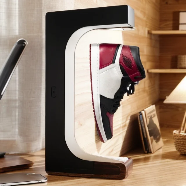 A sneaker in shades of white, black, and red hovers within the Floating Shoe Stand, positioned on a wooden desk beside a lamp and some books.