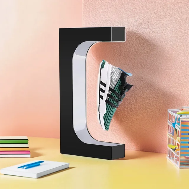 A Floating Shoe Stand holds a sneaker aloft within its black C-shaped magnetic frame, positioned on a desk alongside notebooks, a pen, and a colorful pen holder, all against a two-toned backdrop.