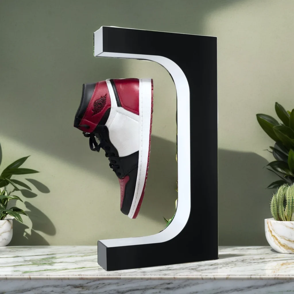 A Floating Shoe Stand in a C-shaped design, featuring a red, black, and white sneaker hovering against its black and white magnetic levitation display on a marble surface, is complemented by potted plants in the backdrop.