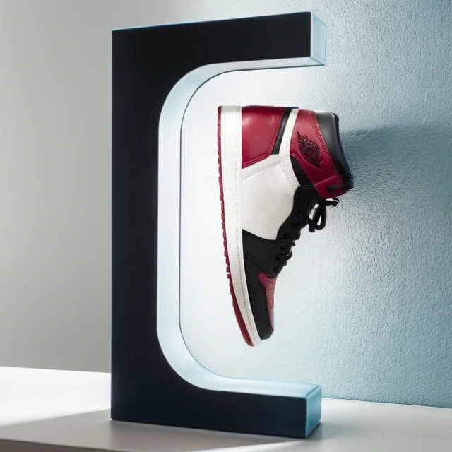 A sneaker in red, white, and black appears to float within a Floating Shoe Stand shaped like a "C," set against a light blue wall.