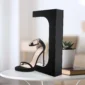 A single black high-heeled shoe displayed on a stand, placed on a white table with a small potted plant and book beside it.