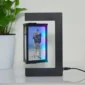 A black photo frame with a floating 3D image of a couple standing on a beach, placed on a white surface beside a green potted plant.