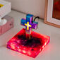 The Floating Cross Nightlight, a vibrant and illuminated Christian cross, stands on a square base featuring a cosmic design. It is placed on a white surface alongside books and a framed picture.