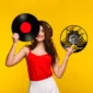Smiling woman in a red top holds a vinyl record and a drum-themed clock against a yellow background, wearing headphones.