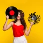 A woman in a red top holds a vinyl record and a music-themed clock. She wears headphones and stands against a yellow background.