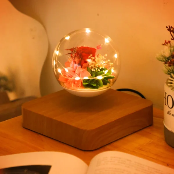 Magnetic Levitating Preserved Flower Rotation Flower Lamp Creative Floating LED Night Light For Home Desk Office Decoration 6 A levitating glass orb with flowers and small lights inside, sits on a wooden base on a table beside an open book.