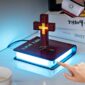A Floating Cross Nightlight designed to resemble a closed book with "Holy Bible" text and an illuminated cross on top, with a finger ready to press the power button.