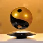 A levitating speaker in the shape of a Yin and Yang symbol, with a reflective gold and black surface, floats above a matching base on a white surface.