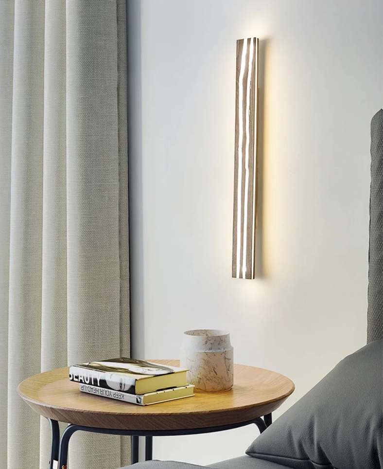 A vertical wall light illuminates a minimalist room with beige curtains, a round wooden table holding books, and a marble candle holder beside a plush chair.