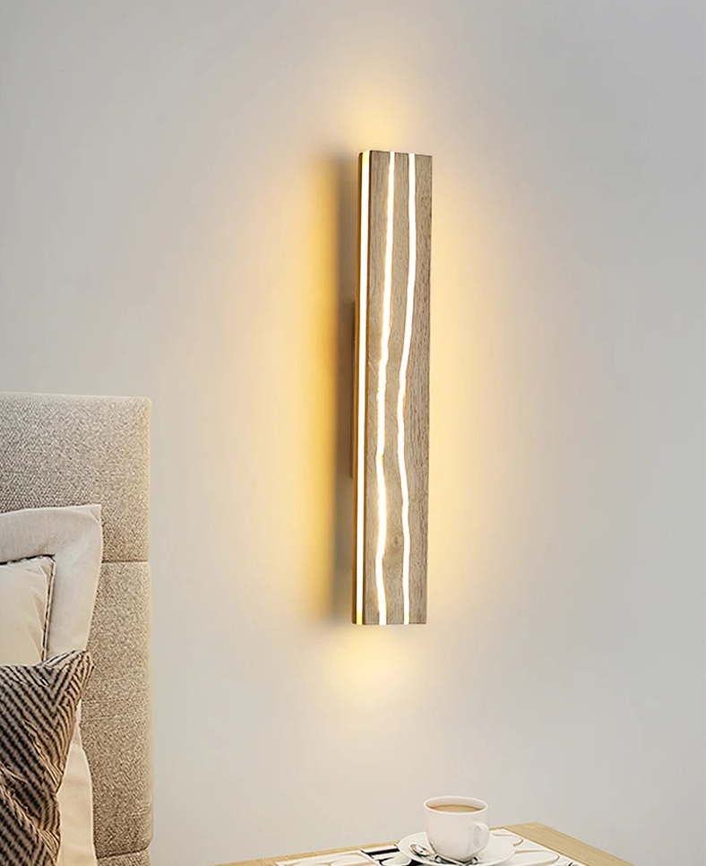 A vertical wooden wall sconce emits warm light next to a bed with beige bedding. A cup and saucer rest on a table beside the bed.