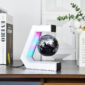 A black and silver globe levitates inside a C-shaped magnetic stand with LED lights, placed on a wooden desk near books and a vase with green and white foliage.