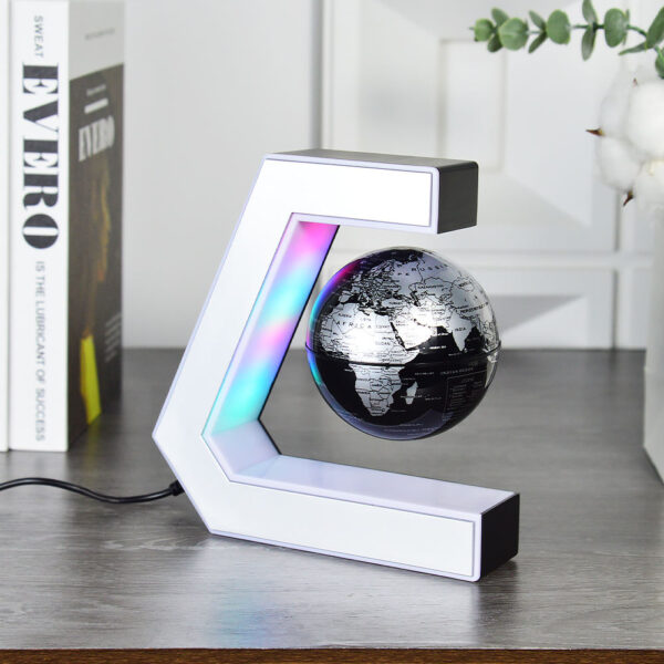 Plastic World Geography Map Rotating Globe Magnetic Levitating Floating Globe for Kids Education Home Office Desktop Decoration 5 A black and silver globe levitates inside a white, C-shaped magnetic stand with LED lights, placed on a wooden desk near a book and decorative items.