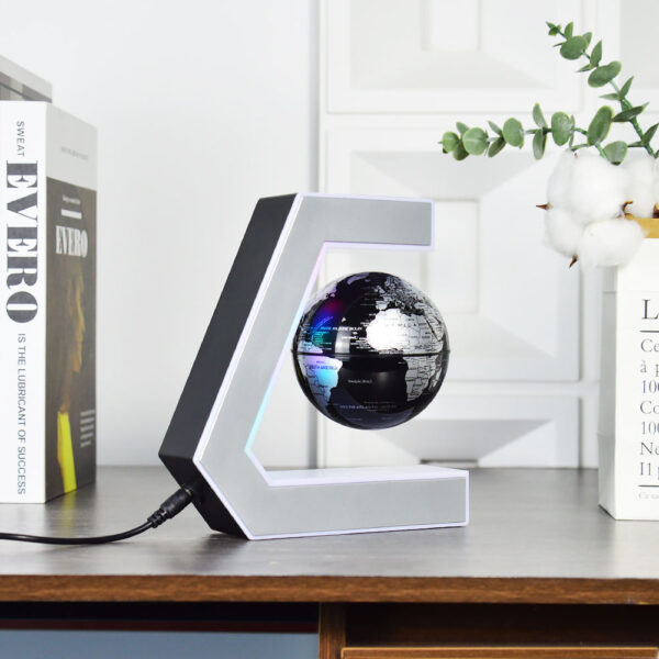 Plastic World Geography Map Rotating Globe Magnetic Levitating Floating Globe for Kids Education Home Office Desktop Decoration 6 A small black floating globe is suspended within a C-shaped magnetic stand on a wooden desk, with books and a plant in the background.