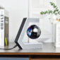 A small black floating globe is suspended within a C-shaped magnetic stand on a wooden desk, with books and a plant in the background.